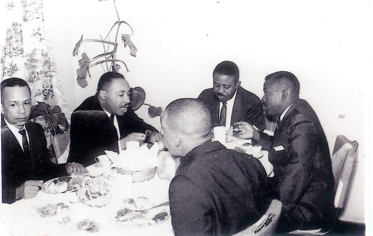 Black-and-white photograph of five men in suits at a dinner table. Dr. Rodgers sits at the far left beside Dr. Martin Luther King Jr.; three other men sit around the table.